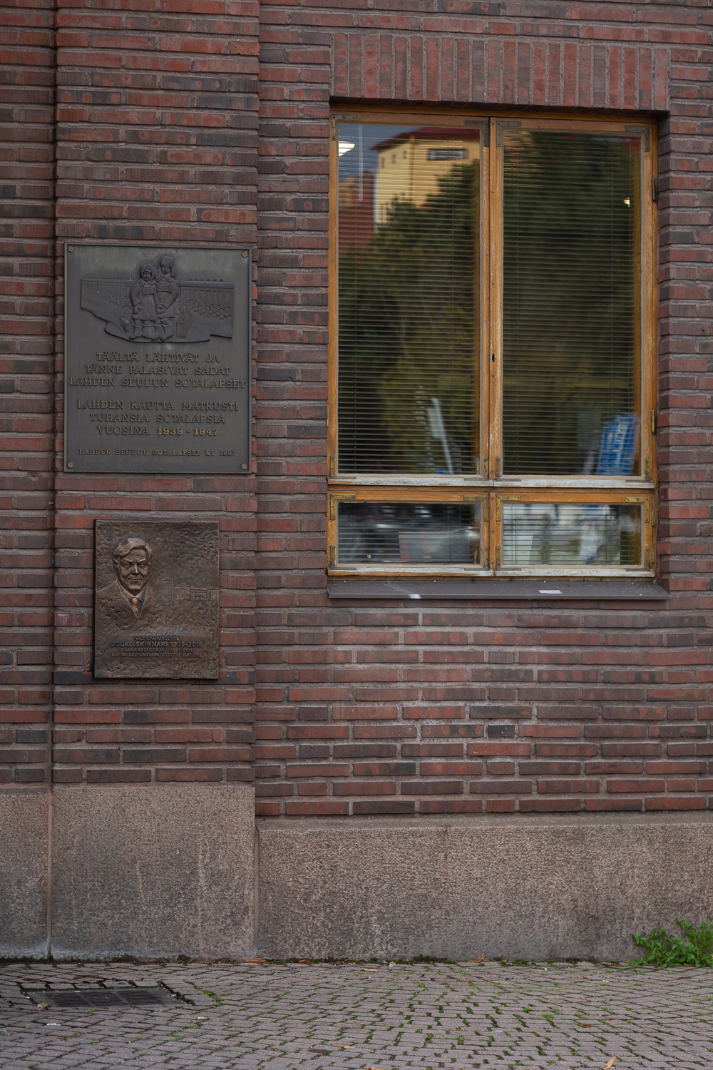 Punatiilisen rakennuksen seinään kiinnitetty reliefi, jossa kaksi lasta laukkuineen junan edessä sekä tekstiä.